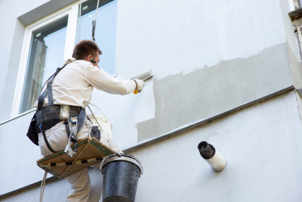 Ein Mann mit Sicherheitsgurt sitzt auf einer Schaukel in der Luft und malt an einer Hauswand die Wand weiß.